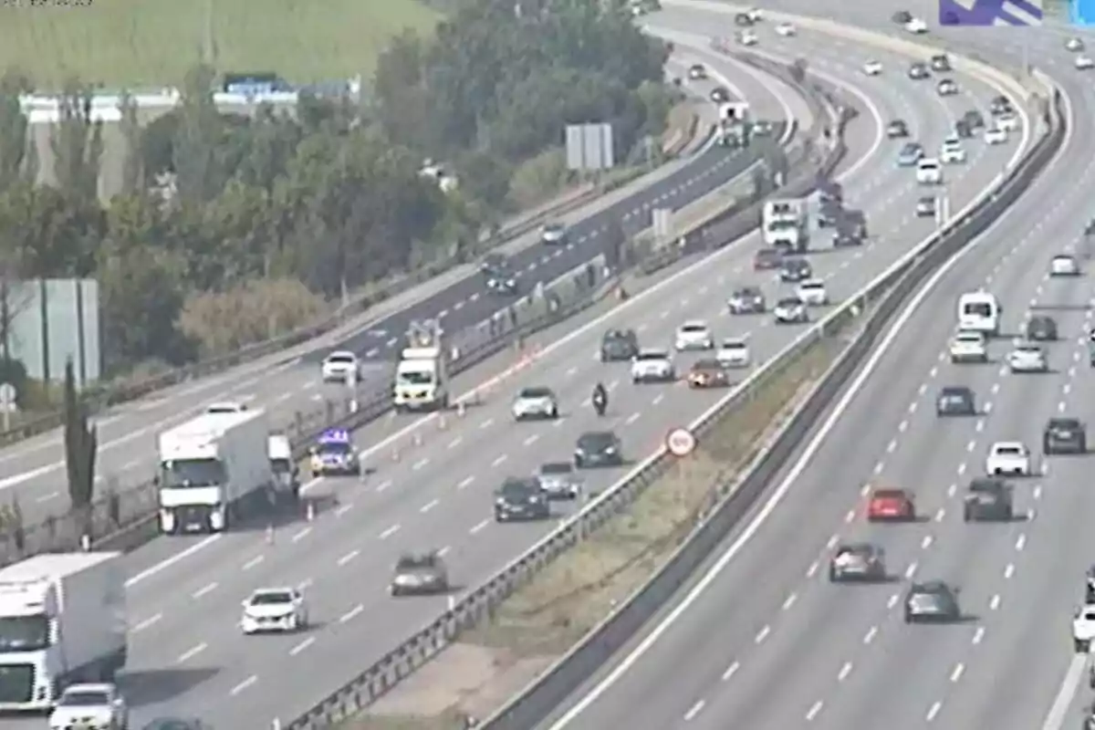Una autopista amb diversos carrils en ambdós sentits mostra trànsit moderat amb vehicles circulant i una àrea de treball senyalitzada amb cons taronja i un vehicle d'emergència amb llums enceses.