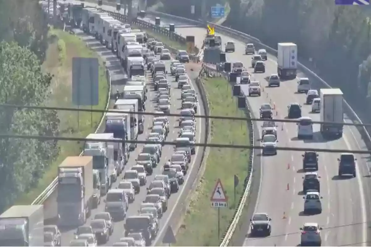 Una carretera con tráfico denso en un carril y tráfico fluido en el otro, con varios vehículos y un cartel de advertencia de obras.