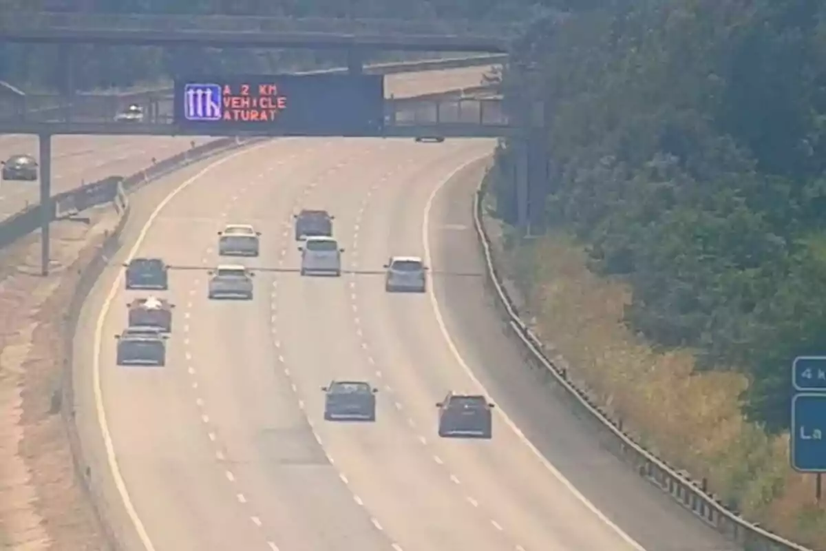 Diversos cotxes circulen per una autopista amb un cartell electrònic que adverteix sobre un vehicle aturat més endavant.