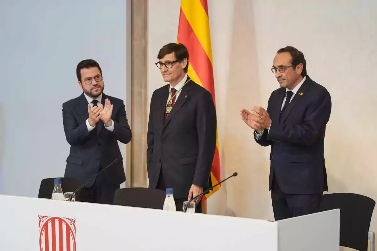 Tres homes de vestit aplaudint en un esdeveniment oficial amb una bandera catalana de fons.