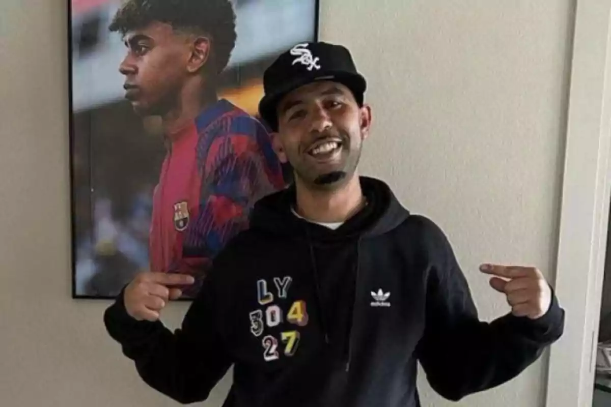 Un hombre sonriente con una gorra negra y una sudadera negra de Adidas, señalando con ambos dedos hacia su sudadera, con un póster de un jugador de fútbol del FC Barcelona en el fondo.