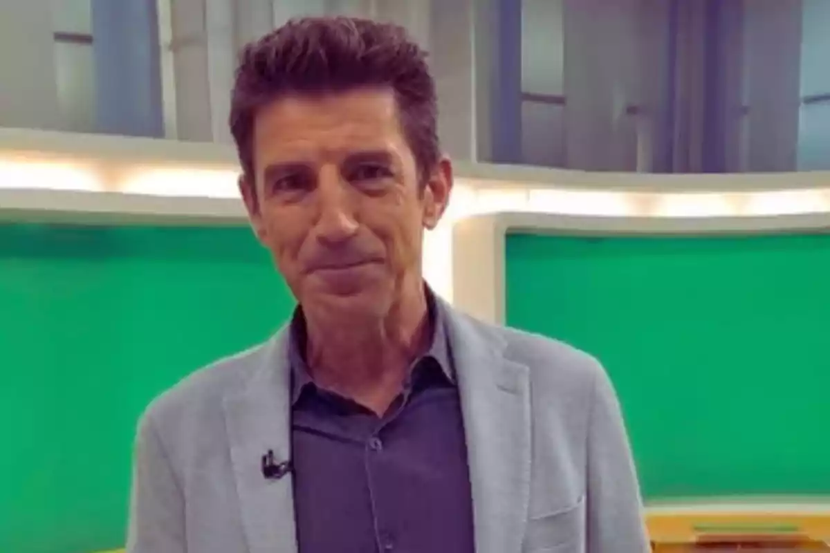 Hombre de pie con chaqueta gris y camisa oscura frente a un fondo verde en un estudio de televisión.