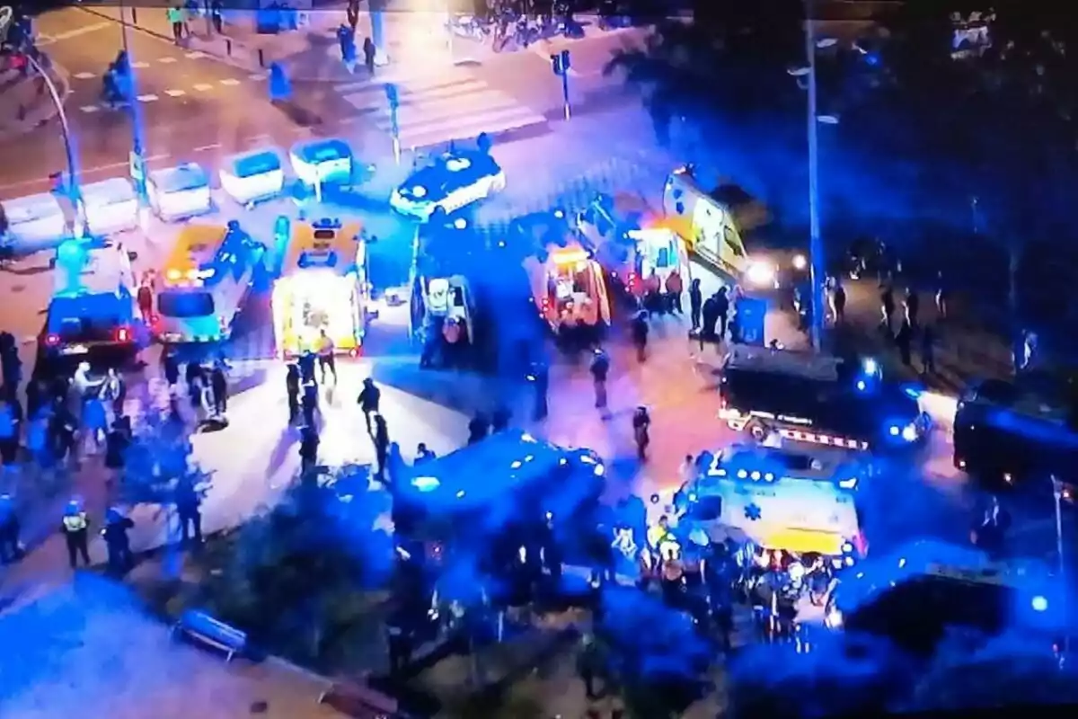 Una escena nocturna mostra una intersecció plena de vehicles d'emergència amb llums blaves i grogues, envoltats de persones i policies, suggerint una situació d'emergència o incident en curs.