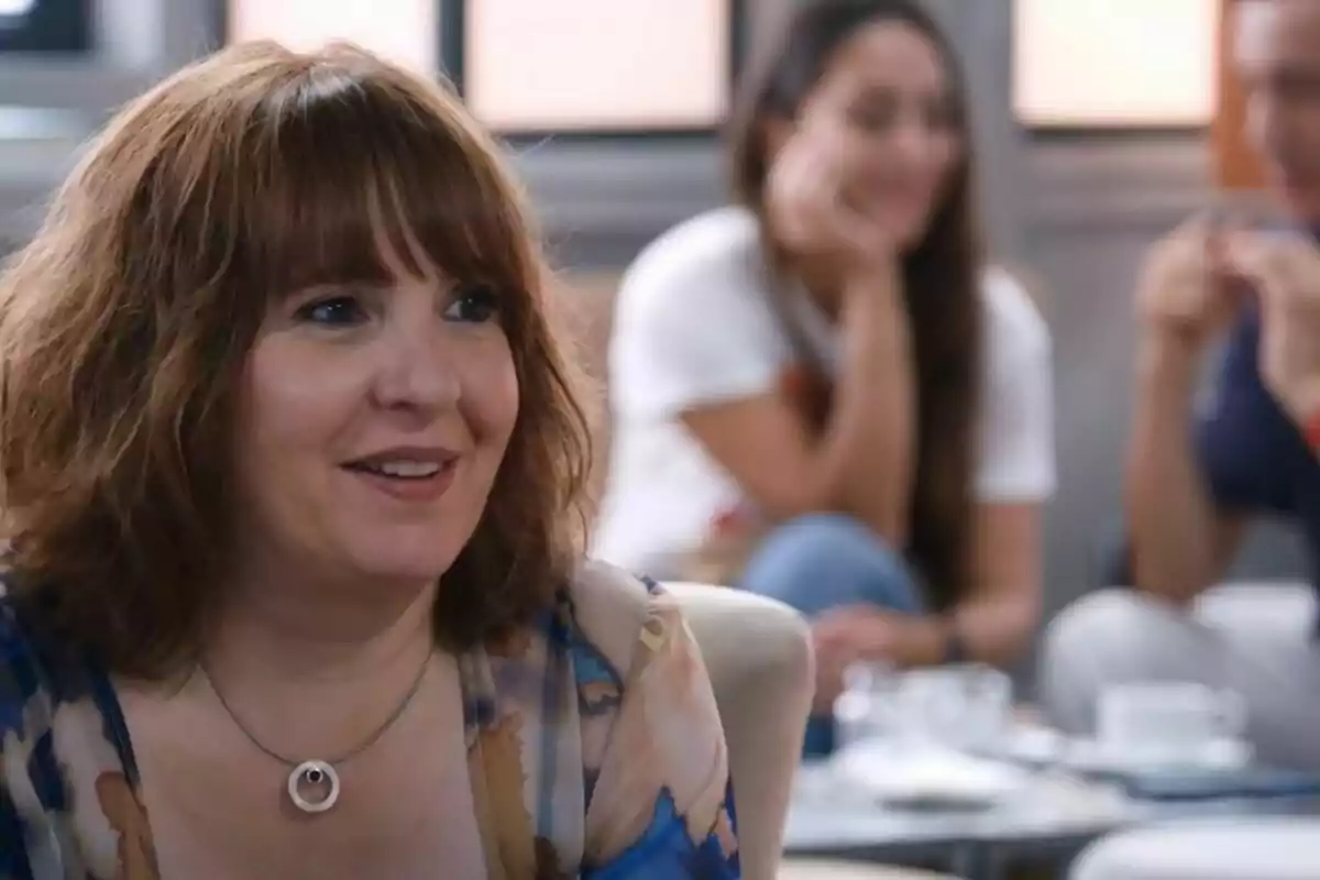 Una mujer sonriente con cabello castaño y un collar está sentada en un ambiente interior con personas desenfocadas al fondo.