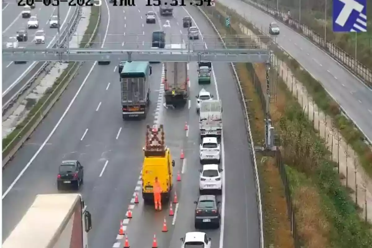 Una carretera amb diversos carrils mostra un vehicle de manteniment groc i un treballador amb armilla taronja col·locant cons taronges per desviar el trànsit, mentre diversos cotxes i camions esperen en una fila.