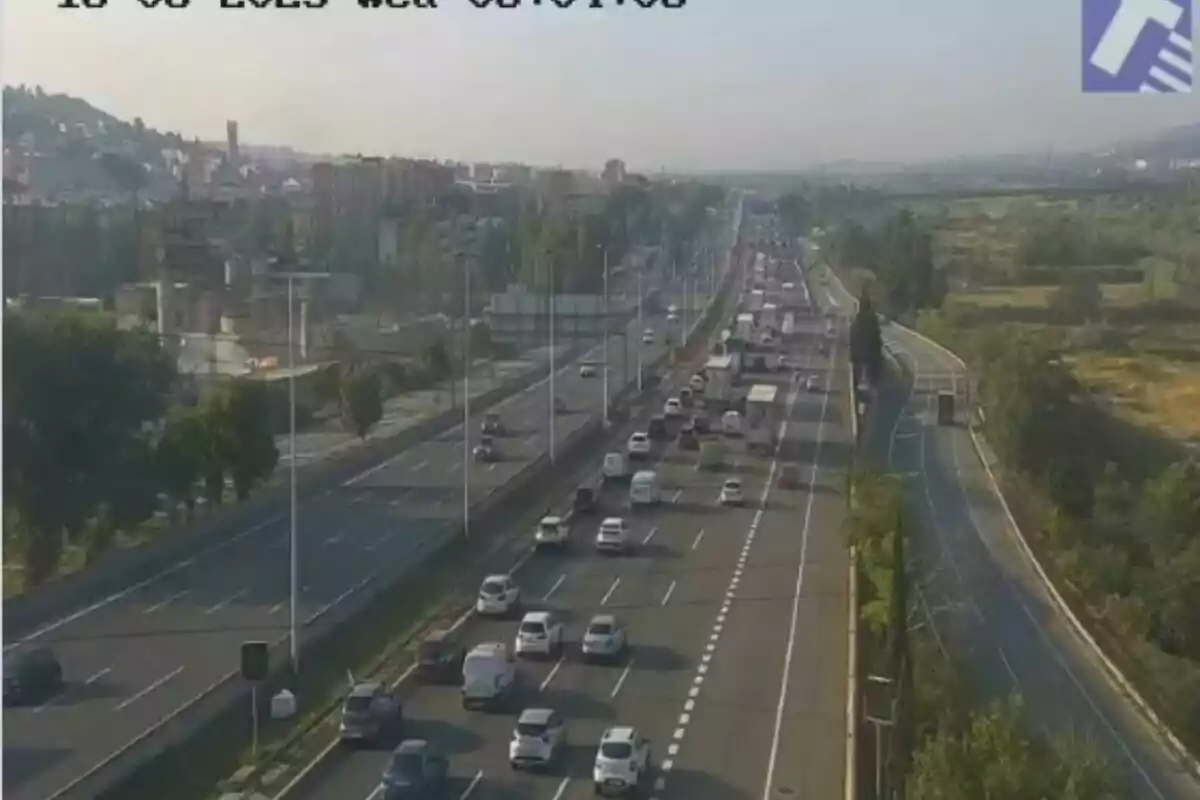 Trànsit moderat en una autopista de diversos carrils envoltada de zones verdes i edificis al fons en un dia assolellat