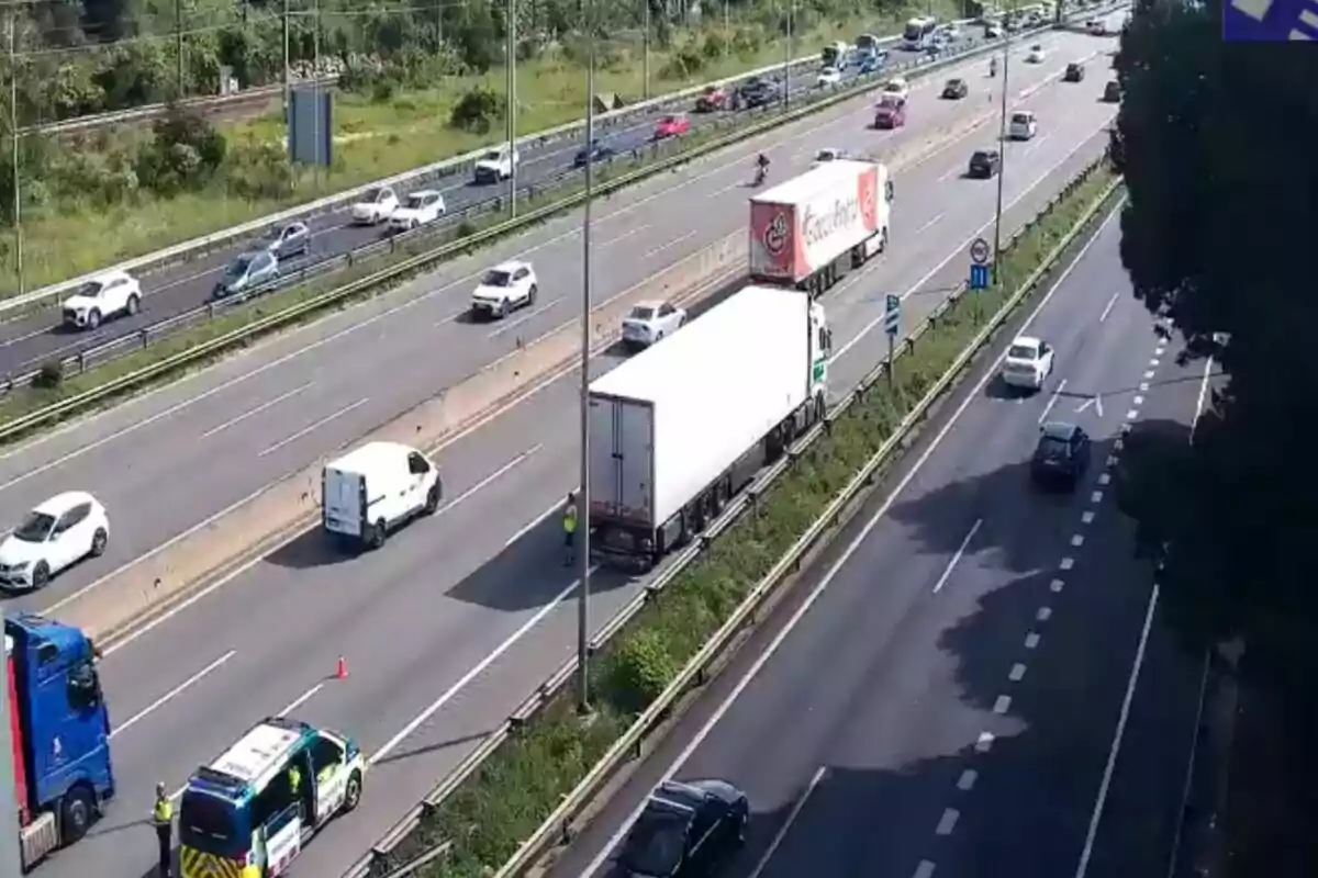 Una carretera amb diversos carrils mostra trànsit de vehicles, incloent camions i cotxes, mentre un vehicle d'emergència està aturat a la vorera amb personal al voltant.