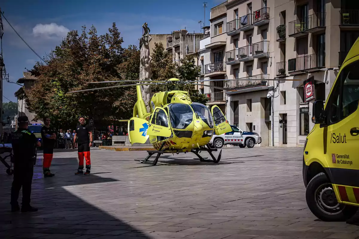 Un helicòpter groc d?emergències mèdiques està estacionat en una plaça, envoltat de personal d?emergència i vehicles de la policia i ambulàncies, amb edificis i arbres al fons.