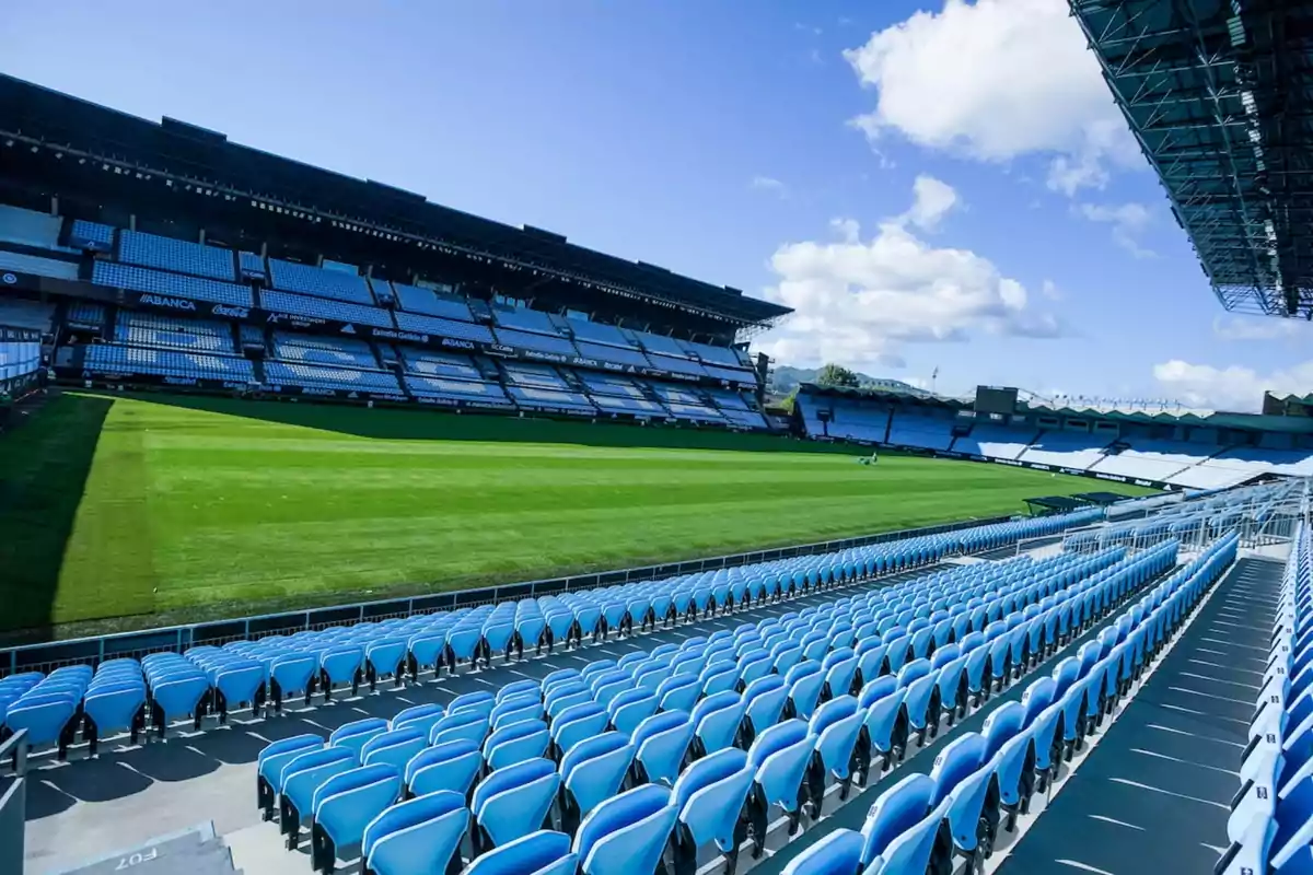 Estadi de futbol buit amb seients blaus i gespa verda sota un cel clar.