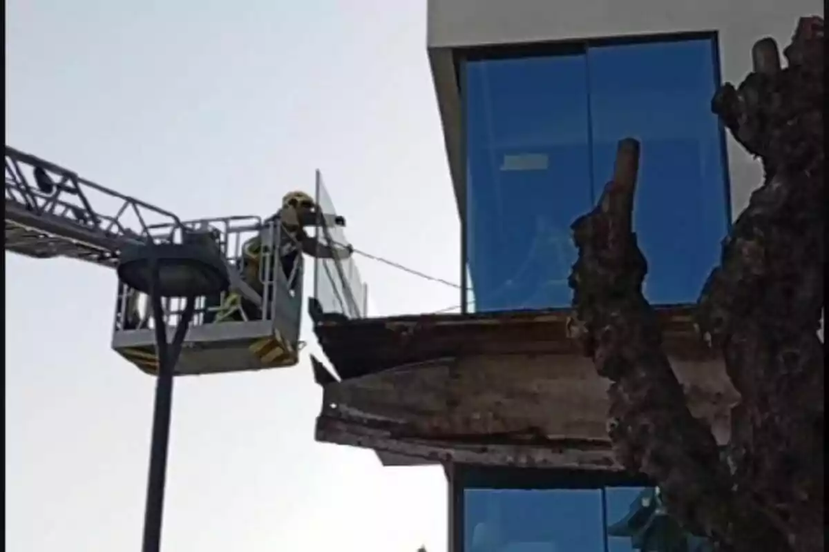 Un bomber en una plataforma elevadora inspecciona un balcó danyat en un edifici modern amb finestres de vidre.