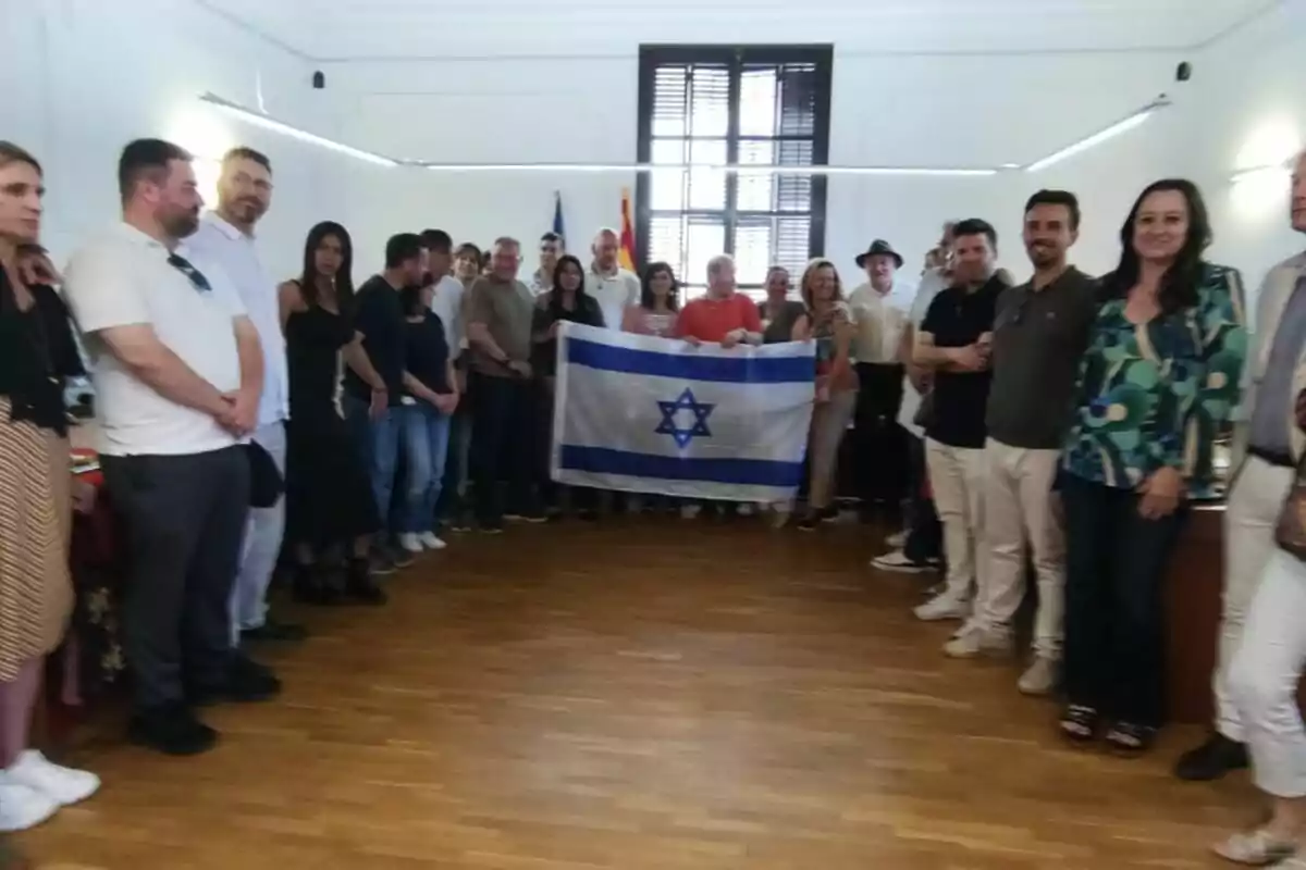 Un grup de persones està reunit en una sala formant un semicercle mentre algunes sostenen una bandera d’Israel al centre. Un grup de persones està reunit en una sala formant un semicercle mentre algunes sostenen una bandera d’Israel al centre.