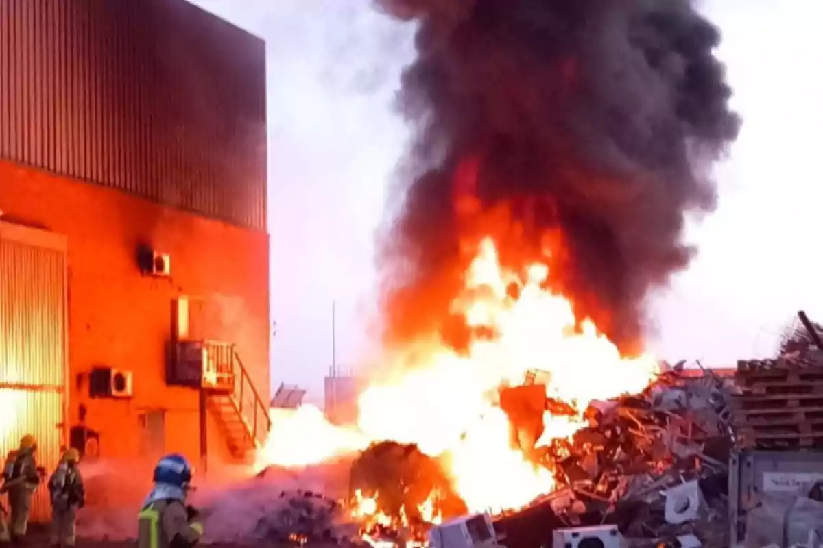 Incendi en un magatzem industrial amb bombers treballant per controlar les flames i una gran columna de fum negre elevant-se cap al cel.