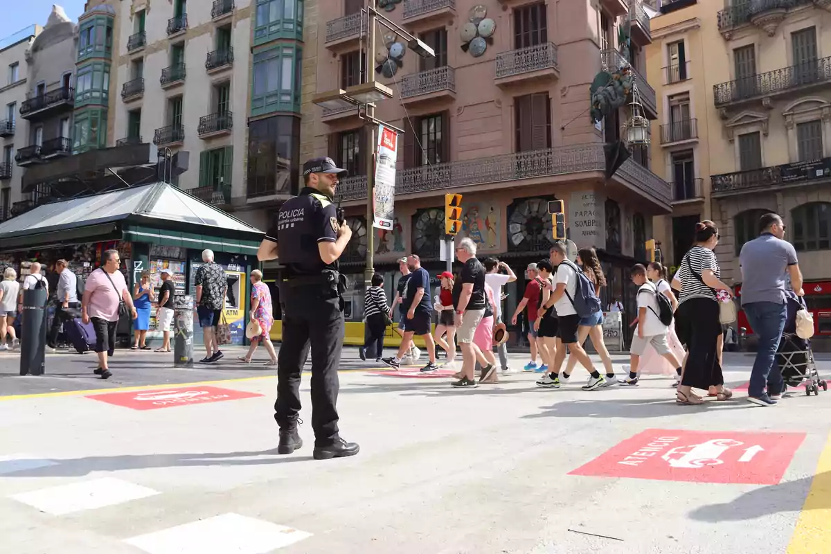 Un policia observa un grup de persones creuant un carrer en una zona urbana amb edificis antics i un quiosc al fons.