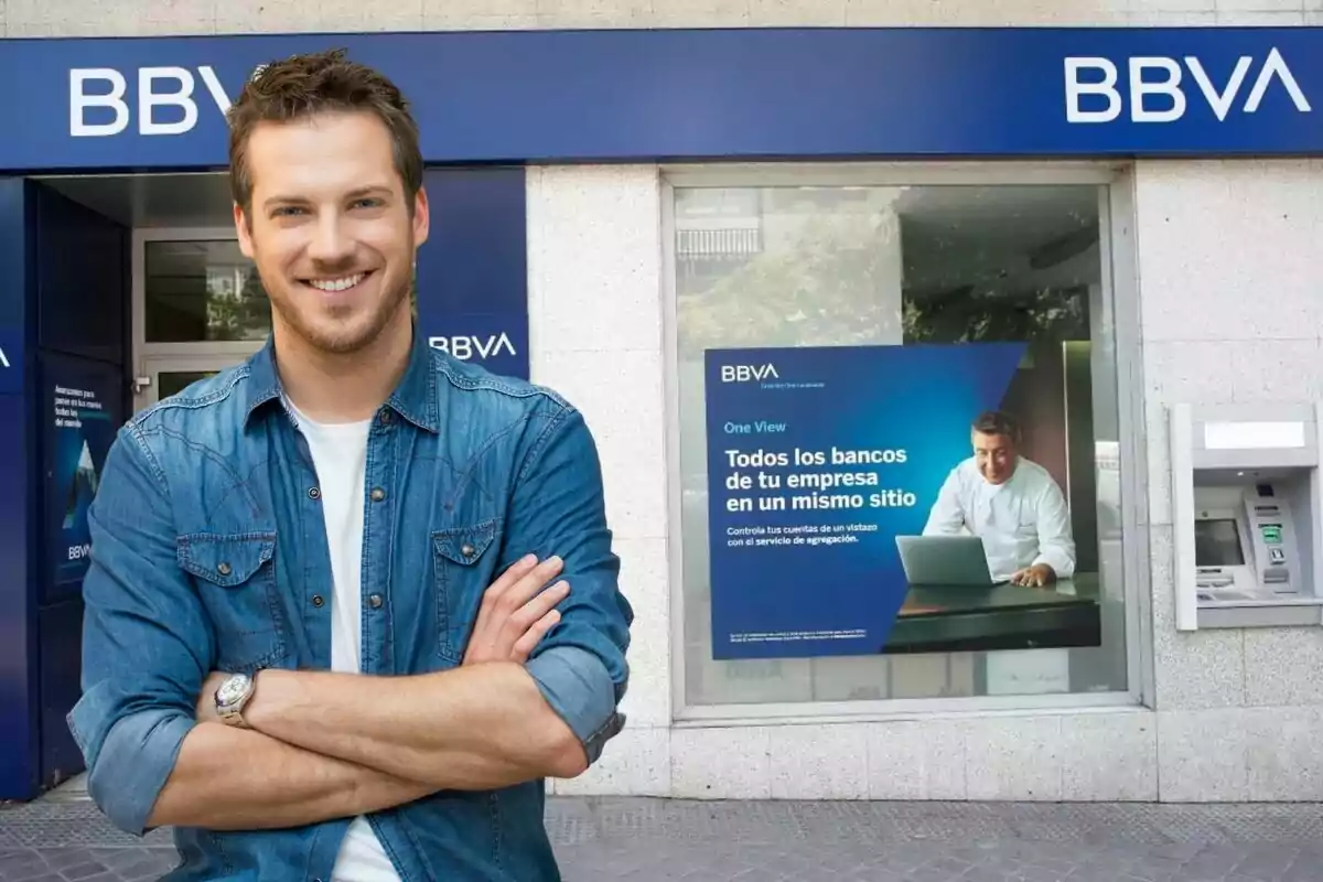 Un hombre sonriente con camisa de mezclilla está de pie frente a una sucursal de BBVA.