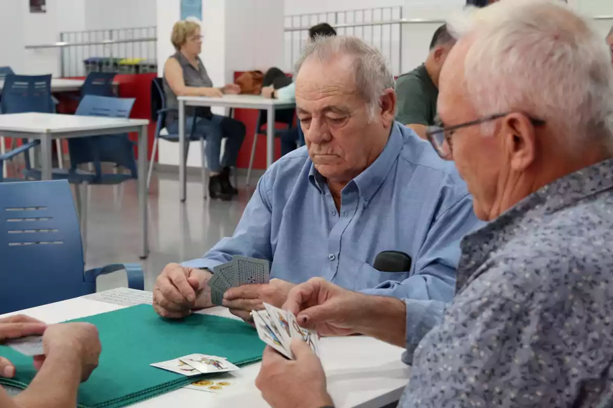 Un grupo de personas mayores juega a las cartas en una sala con mesas y sillas.