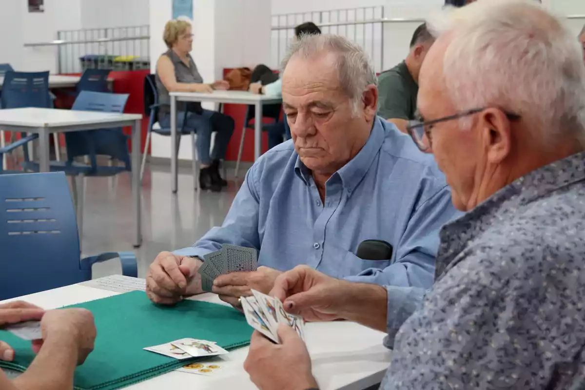 Un grup de gent gran juga a cartes en una sala amb taules i cadires blaves.