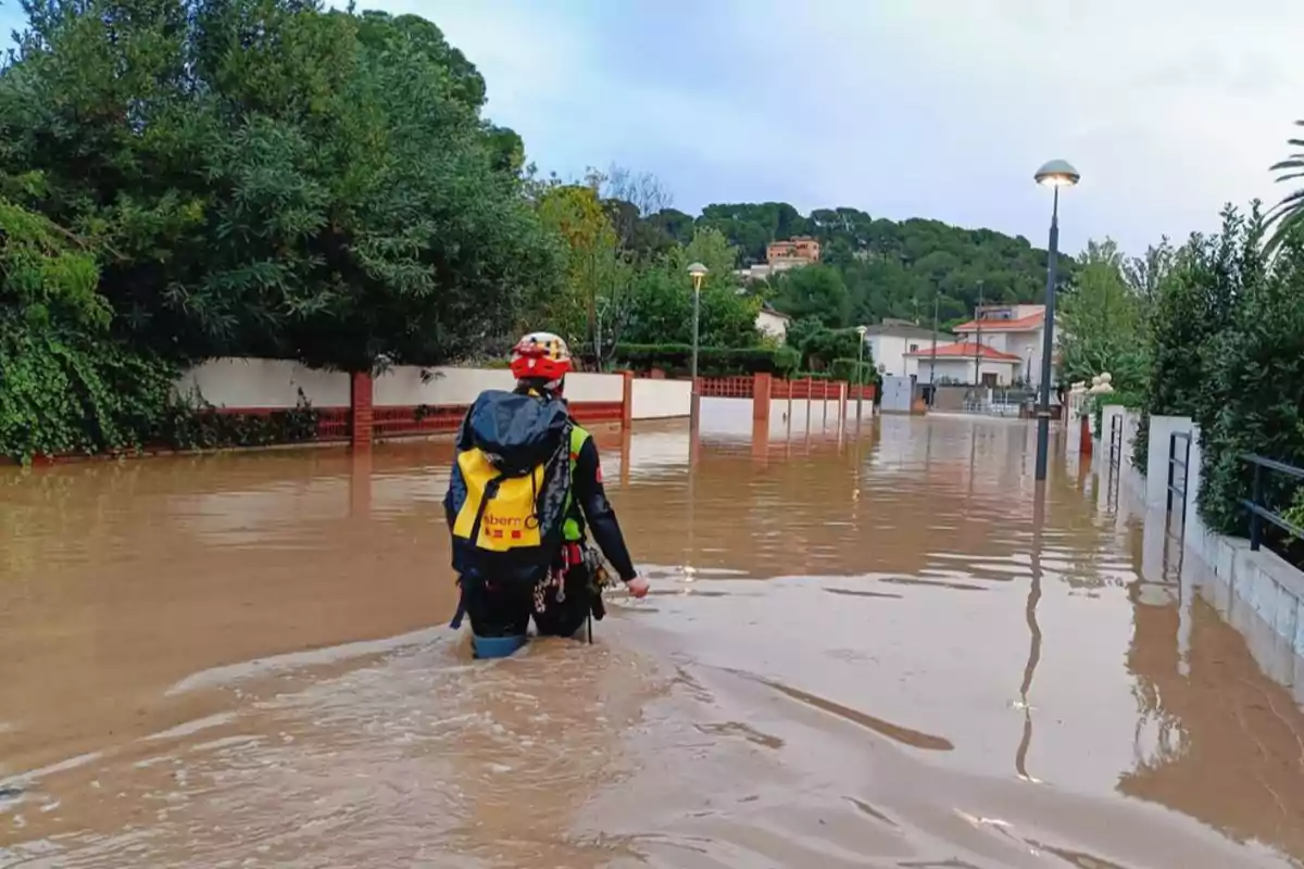 Una persona con equipo de rescate camina por una calle inundada rodeada de árboles y casas.