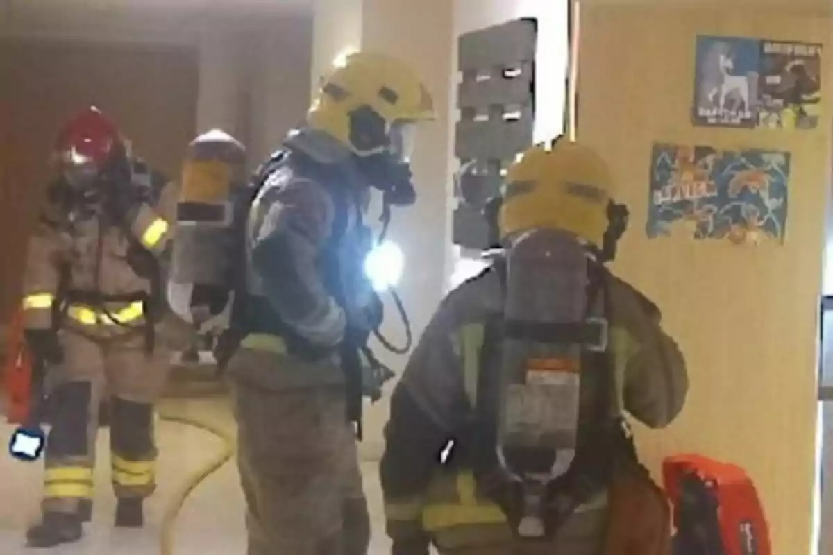 Tres bombers amb equip de protecció i ampolles d’oxigen revisen l’interior d’un edifici mentre una mànega groga s’estén pel terra i hi ha cartells a la paret.