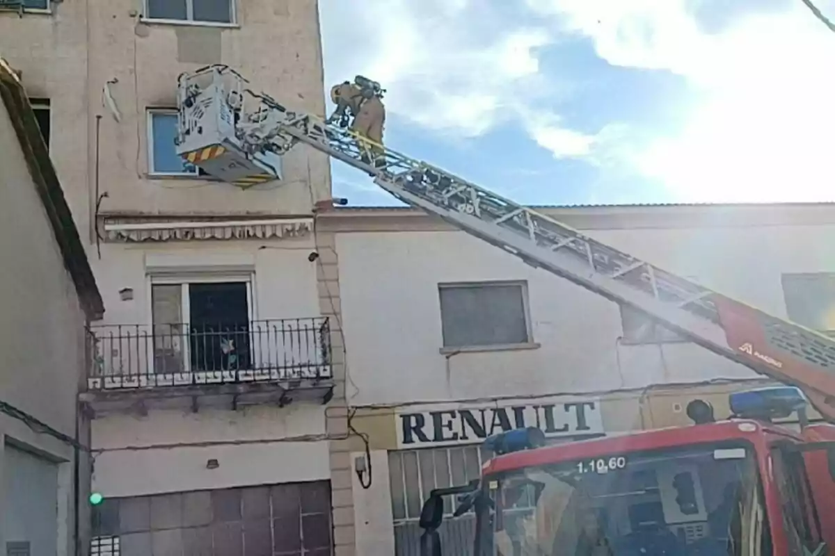 Un camió de bombers amb una escala estesa és davant d'un edifici, mentre un bomber treballa en una plataforma elevada a prop d'una finestra.