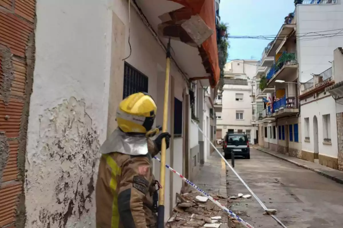 Un bomber amb casc groc inspecciona un edifici en un carrer estret mentre sosté una eina llarga, amb runes a terra i una cinta de seguretat delimitant l'àrea.