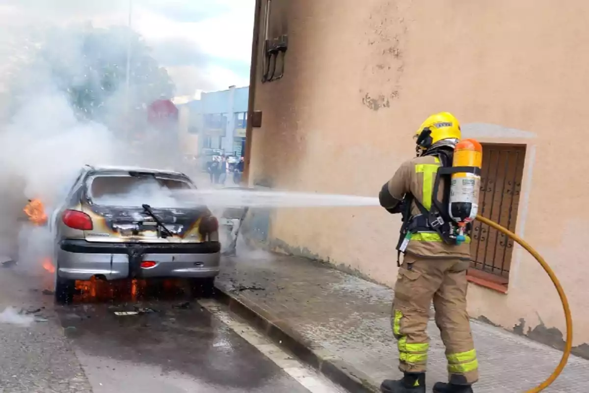 Un bomber apaga un incendi en un automòbil al carrer fent servir una mànega d'aigua.