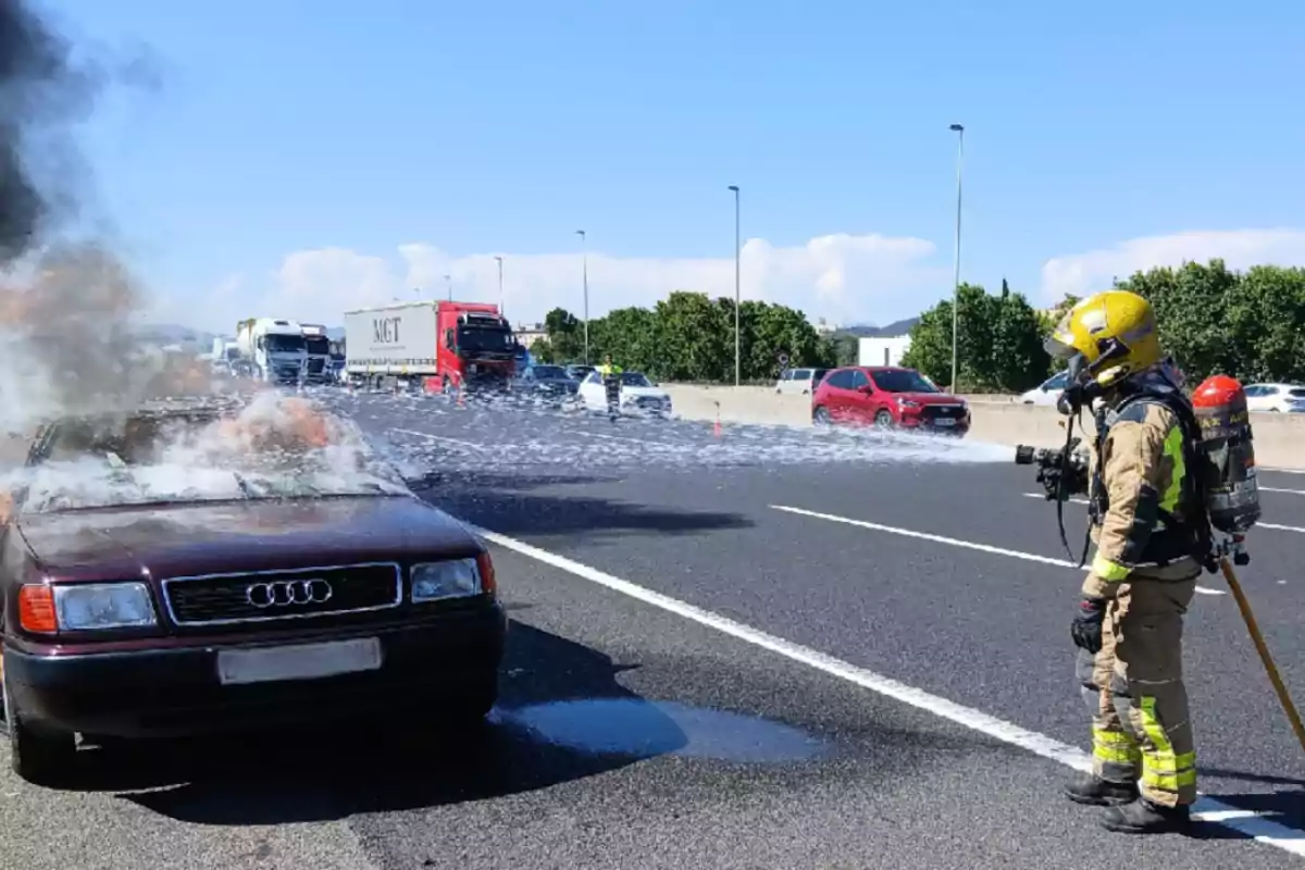 Un bomber apaga l’incendi d’un cotxe en una autopista mentre diversos vehicles passen pel carril contrari.