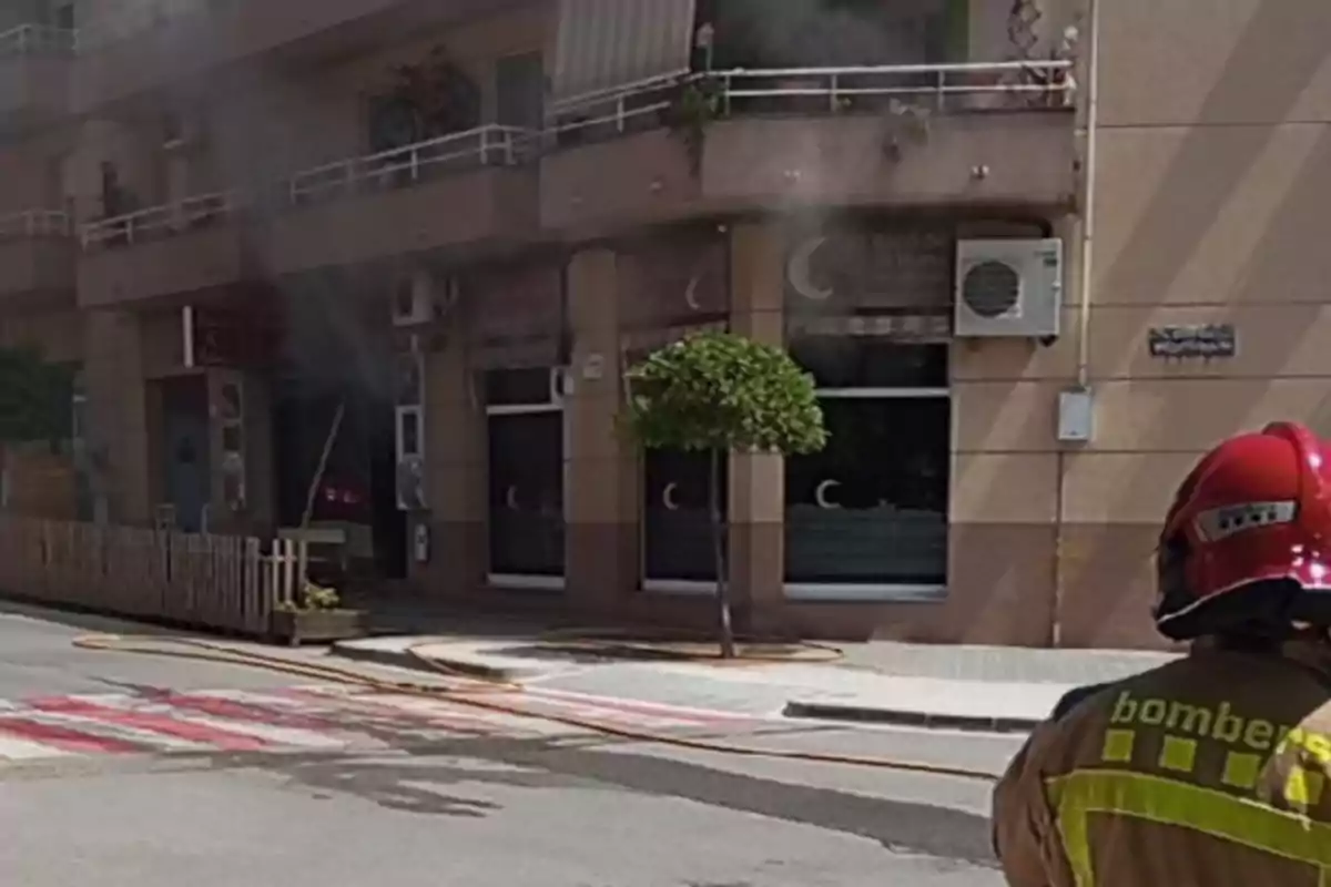 Un bomber amb casc vermell i uniforme observa un edifici del qual surt fum, amb mànegues esteses al carrer.