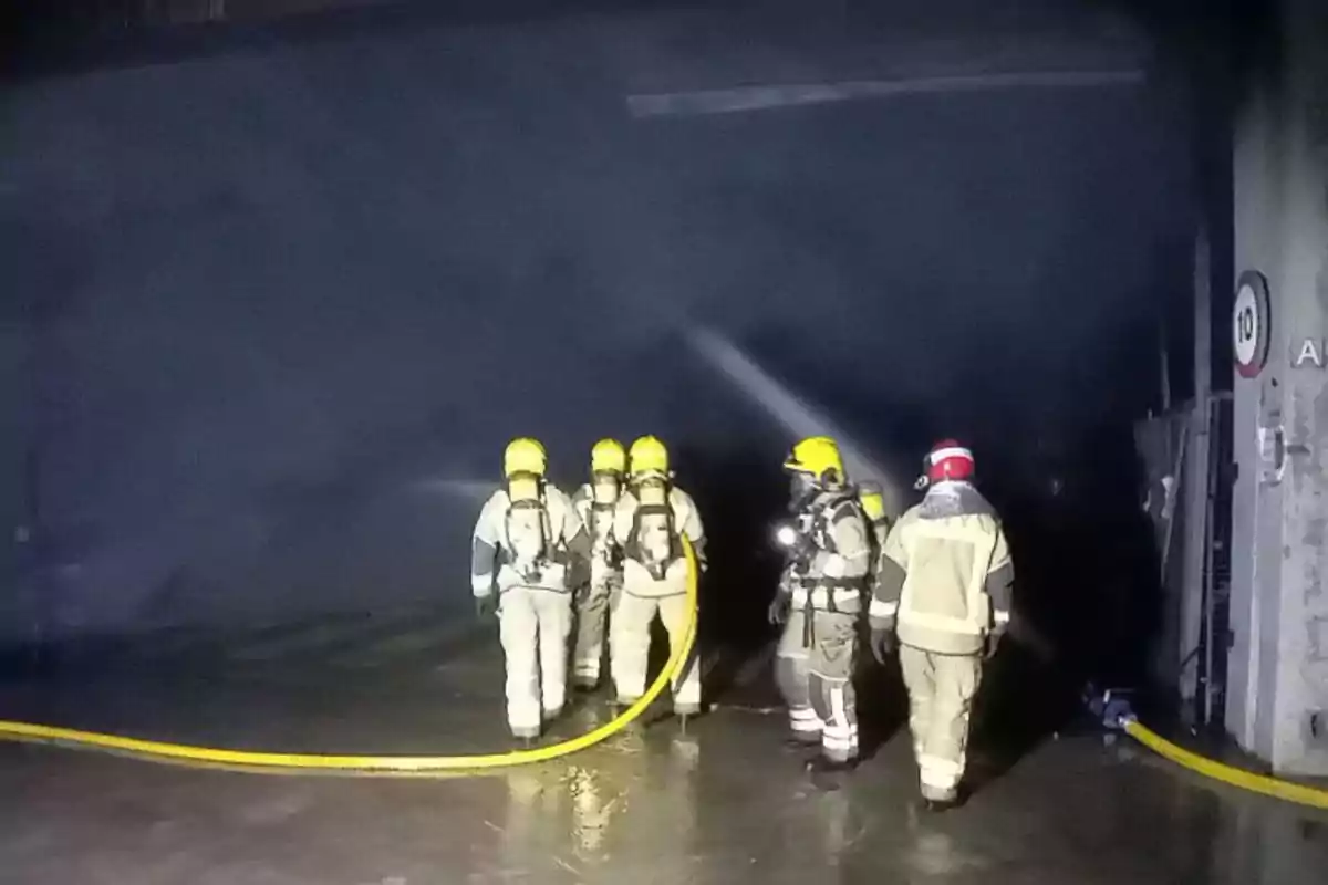 Un grupo de bomberos con equipo de protección y cascos amarillos sostiene una manguera mientras trabaja en un área oscura.