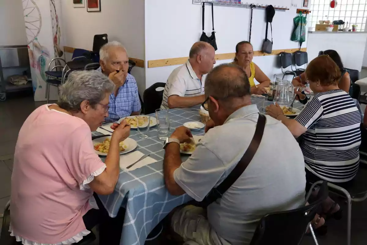 Un grup de persones grans compartint un dinar en una taula amb estovalles blaves a quadres en un menjador.