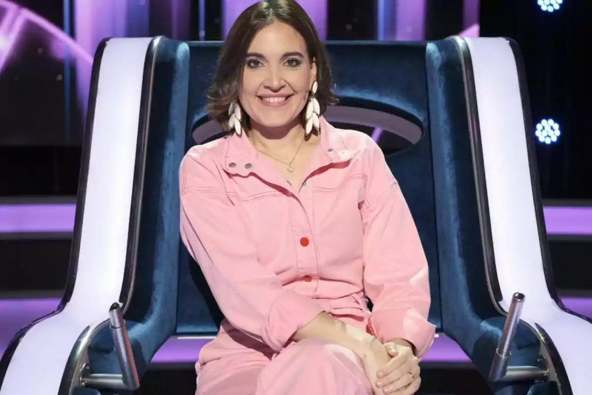 Una mujer sonriente con un mono rosa y pendientes grandes está sentada en una silla moderna en un entorno iluminado con luces púrpuras.