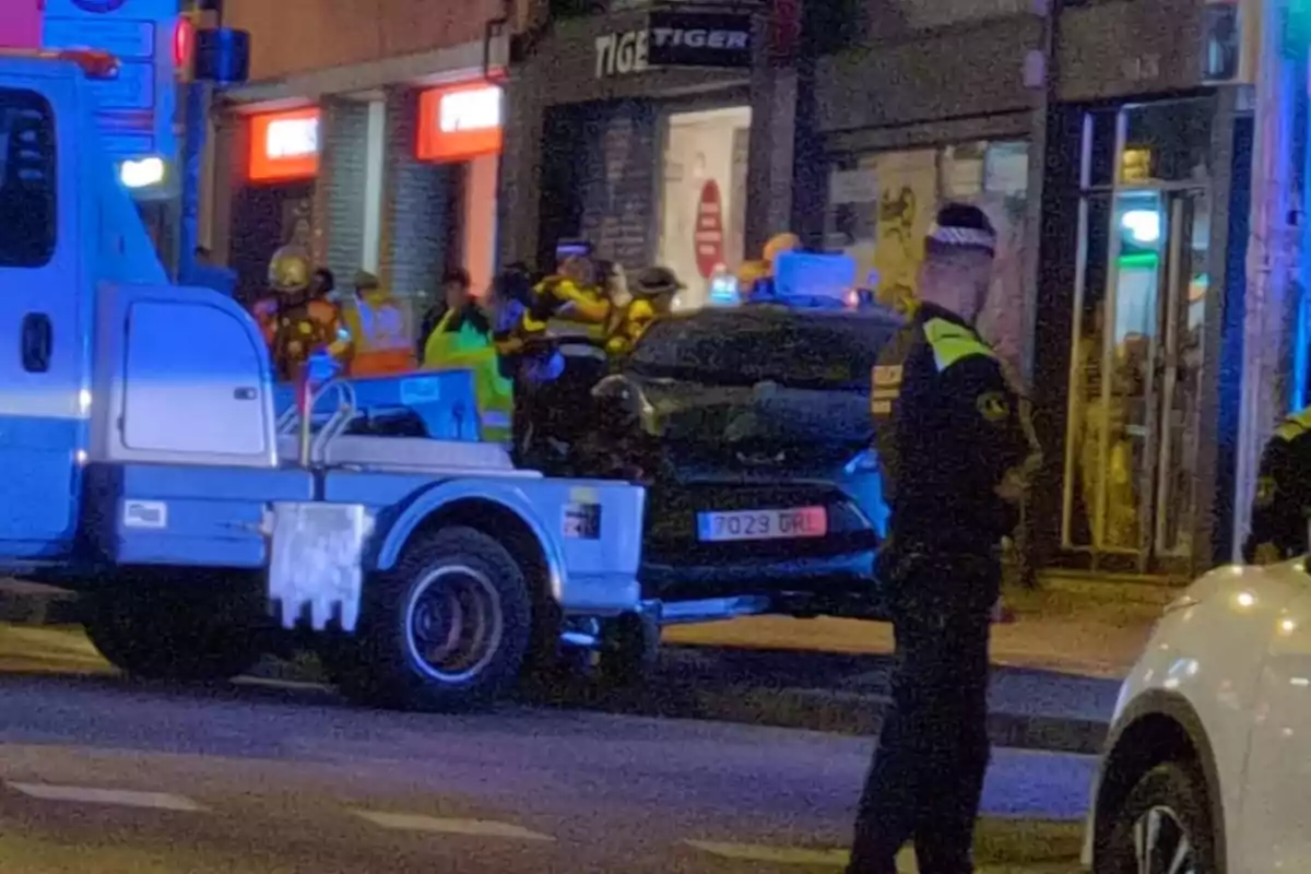 Una grua remolca un cotxe negre mentre que diversos policies i personal d'emergència són presents en un carrer il·luminat per llums d'emergència.