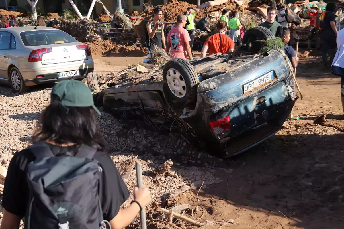 Una escena de desastre natural muestra un coche volcado y cubierto de lodo rodeado de personas que observan y caminan por el área afectada.