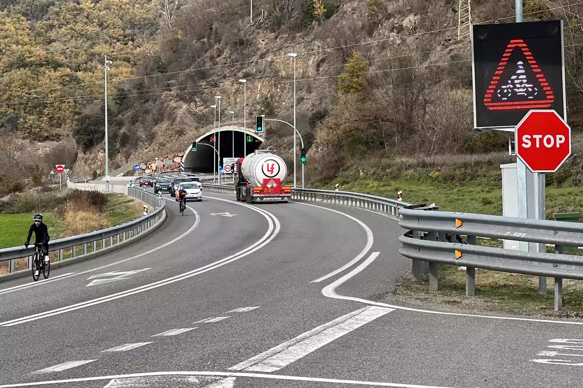 Una carretera amb ciclistes, cotxes i un camió cisterna que es dirigeix cap a un túnel en una zona muntanyosa, amb senyals de trànsit visibles, incloent-hi un senyal de stop i un senyal lluminós d'advertiment. Una carretera amb ciclistes, cotxes i un camió cisterna que es dirigeix cap a un túnel en una zona muntanyosa, amb senyals de trànsit visibles, incloent-hi un senyal de stop i un senyal lluminós d'advertiment.