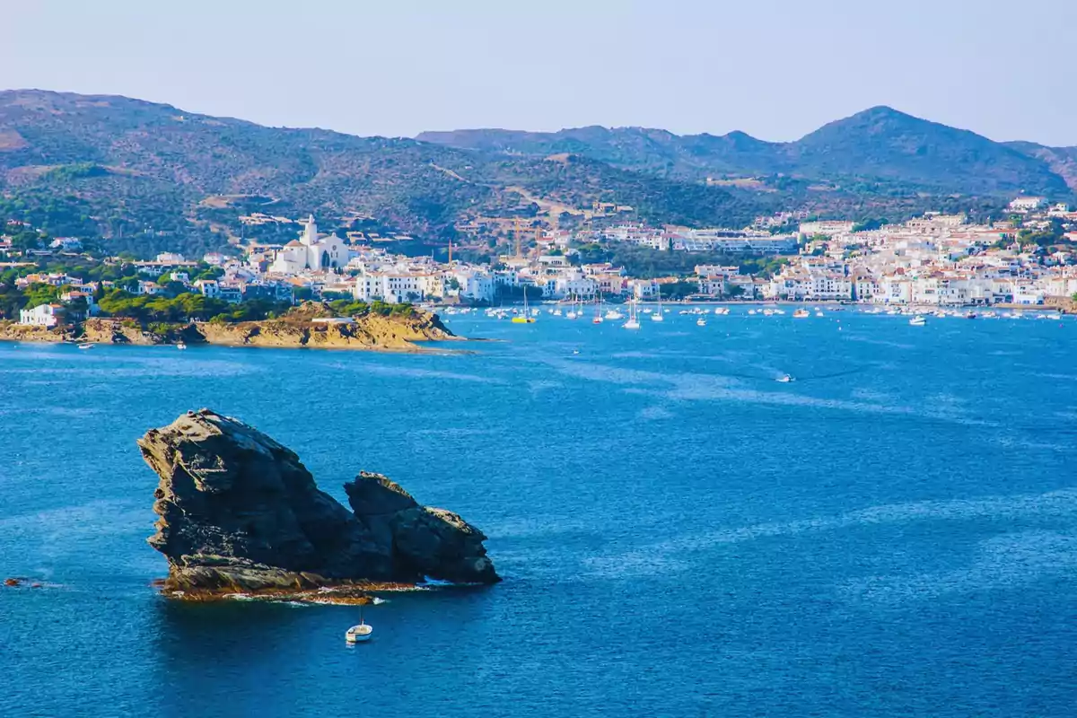 Vista panoràmica d´un poble costaner amb un mar blau i una formació rocosa en primer pla.