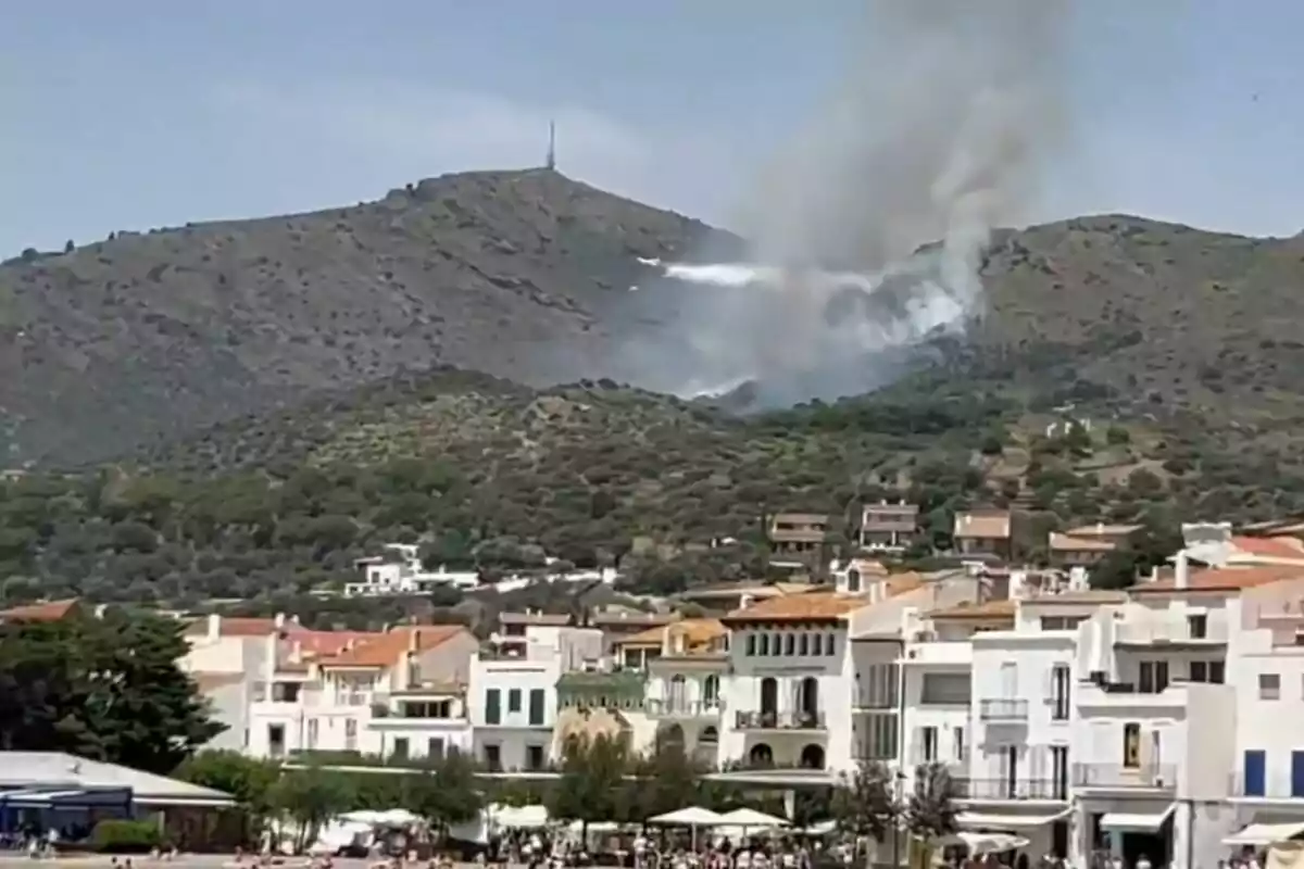 Fum sortint d’un incendi forestal en un vessant prop d’un poble amb cases blanques i teulades de teula vermella