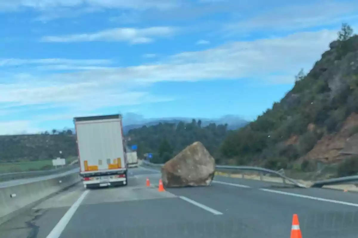 Un camió aturat en una carretera a causa d'una gran roca que bloqueja el pas, amb cons taronja senyalitzant l'obstacle.