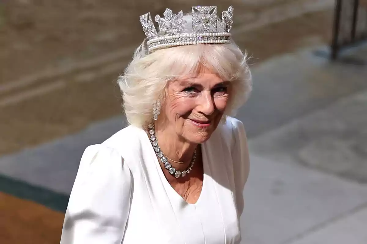 Queen Camilla with blonde hair and a tiara smiles while wearing an elegant white dress. Queen Camilla with blonde hair and a tiara smiles while wearing an elegant white dress.