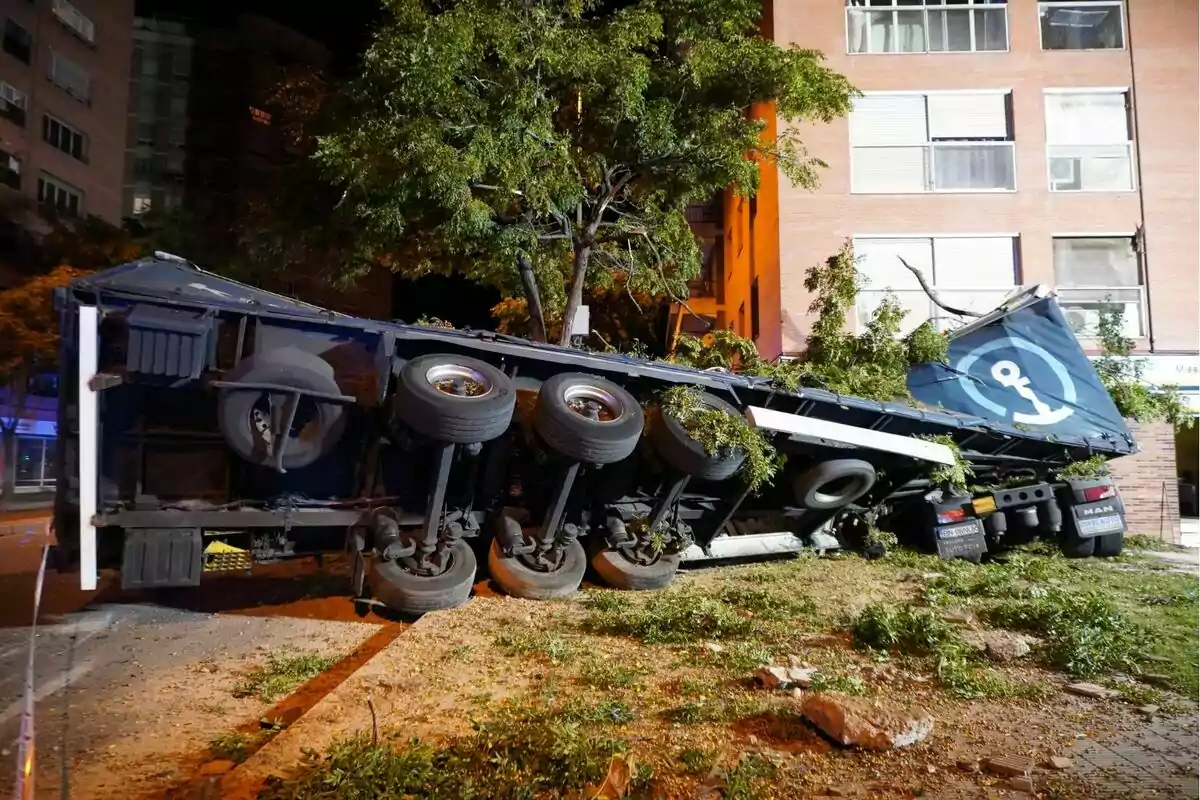 Un camión volcado en una calle de la ciudad, con árboles y edificios de fondo.