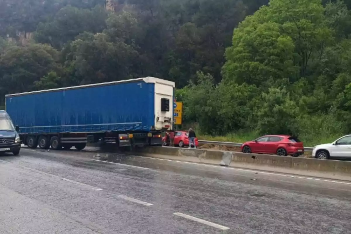 Un camió blau està travessat a la carretera, bloquejant diversos carrils, mentre que diversos cotxes estan aturats a prop de la mitjana en un entorn envoltat d'arbres.