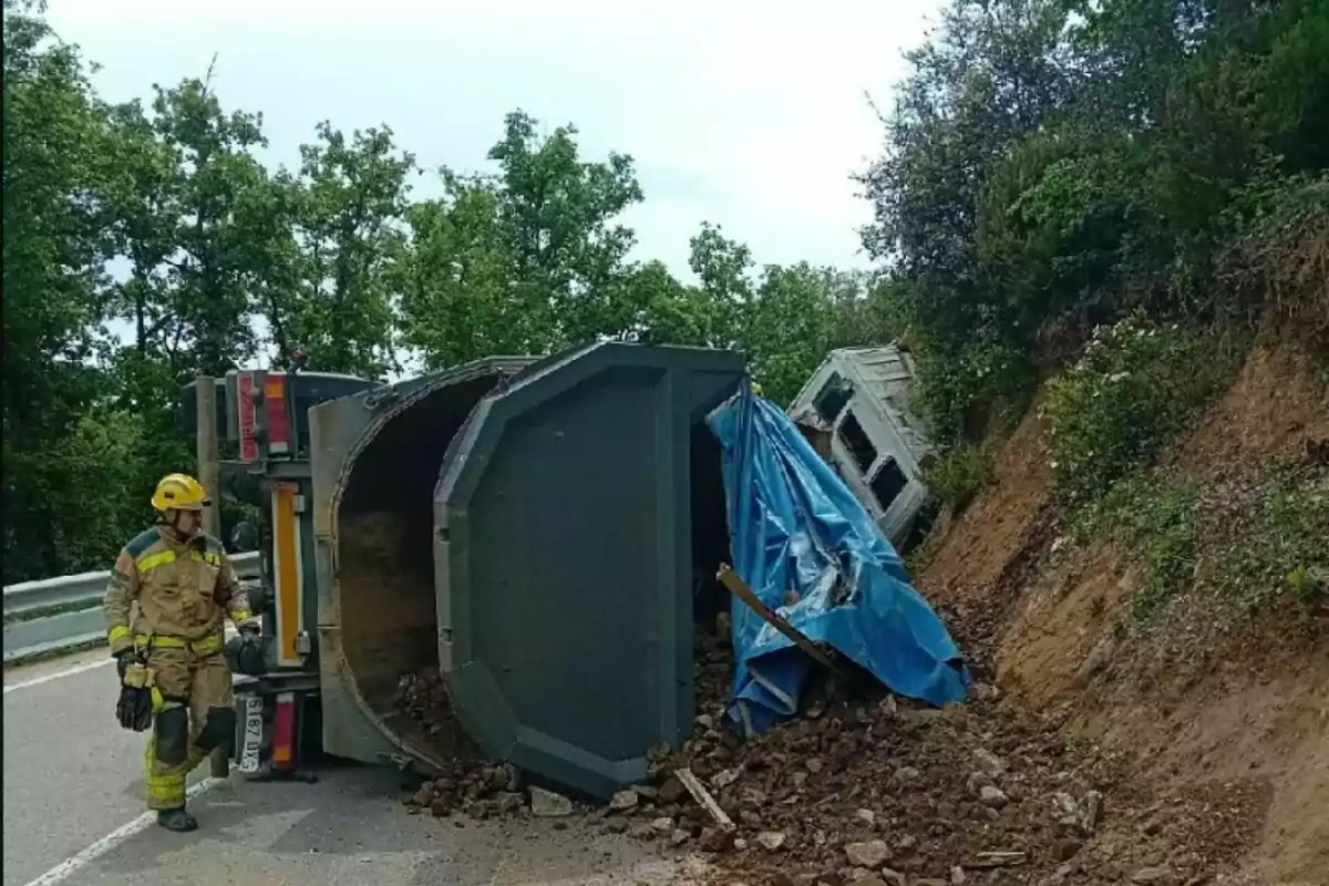 Un camió bolcat en una carretera amb un bomber inspeccionant l'escena i runa escampada al voltant.