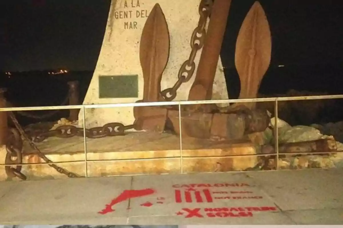 Una gran àncora rovellada està recolzada en un monument de pedra amb una inscripció que diu “A LA GENT DEL MAR” ia terra hi ha un grafit vermell amb la paraula “CATALÒNIA” i altres textos.