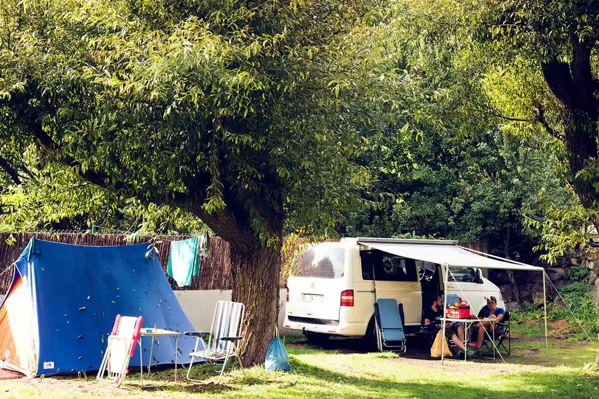 Una escena de campament amb una tenda de campanya blava i una furgoneta blanca amb un tendal desplegat, envoltada d'arbres i vegetació, amb persones assegudes a cadires plegables gaudint de l'entorn.