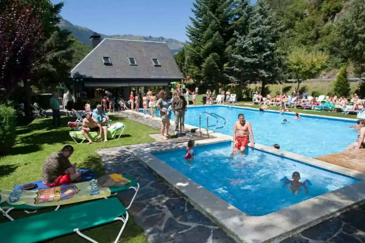 Persones gaudint d'una piscina exterior en un entorn muntanyós amb arbres i gespa, algunes persones neden mentre altres descansen en gandules.
