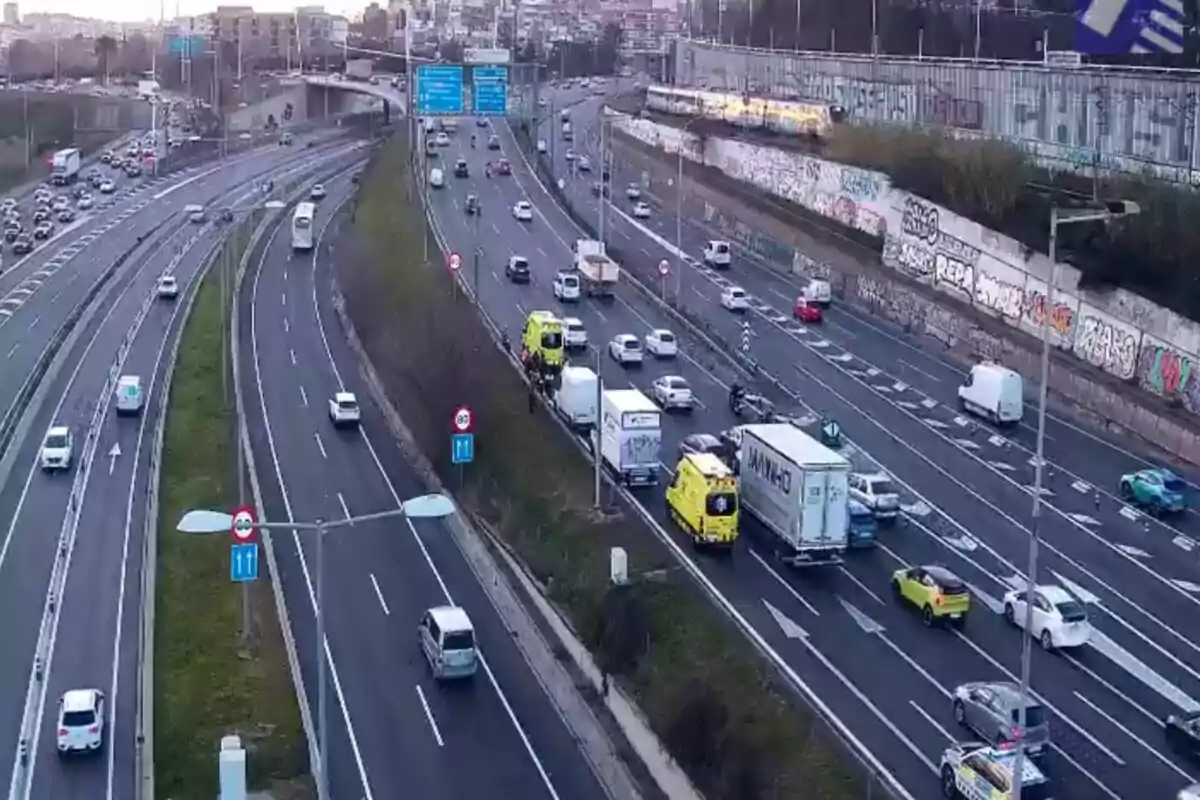 Una autopista amb diversos carrils mostra trànsit moderat, incloent cotxes, camions i una ambulància, mentre que al fons s'observen edificis i senyals de trànsit.