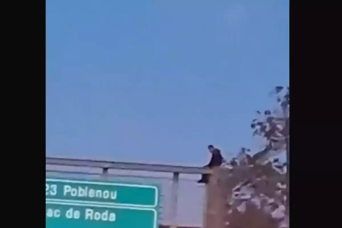 Una persona asseguda a la vora d'un pont amb un cartell verd que indica adreces cap al Poblenou i el Bac de Roda.