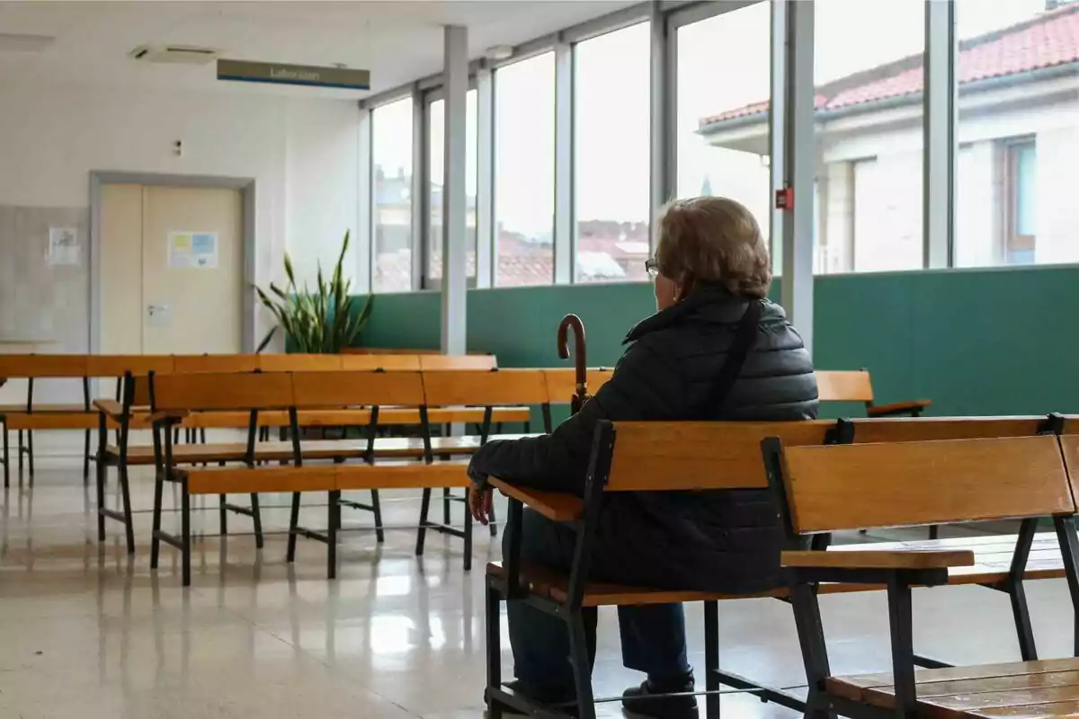 Una persona asseguda en una sala d'espera buida amb bancs de fusta i un bastó a la mà.