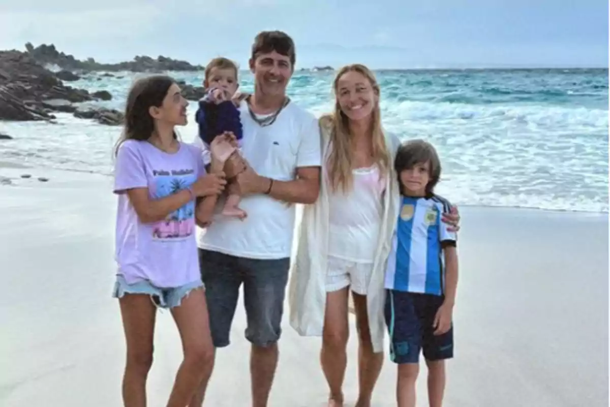 Una família somrient posant a la platja amb el mar i roques de fons.