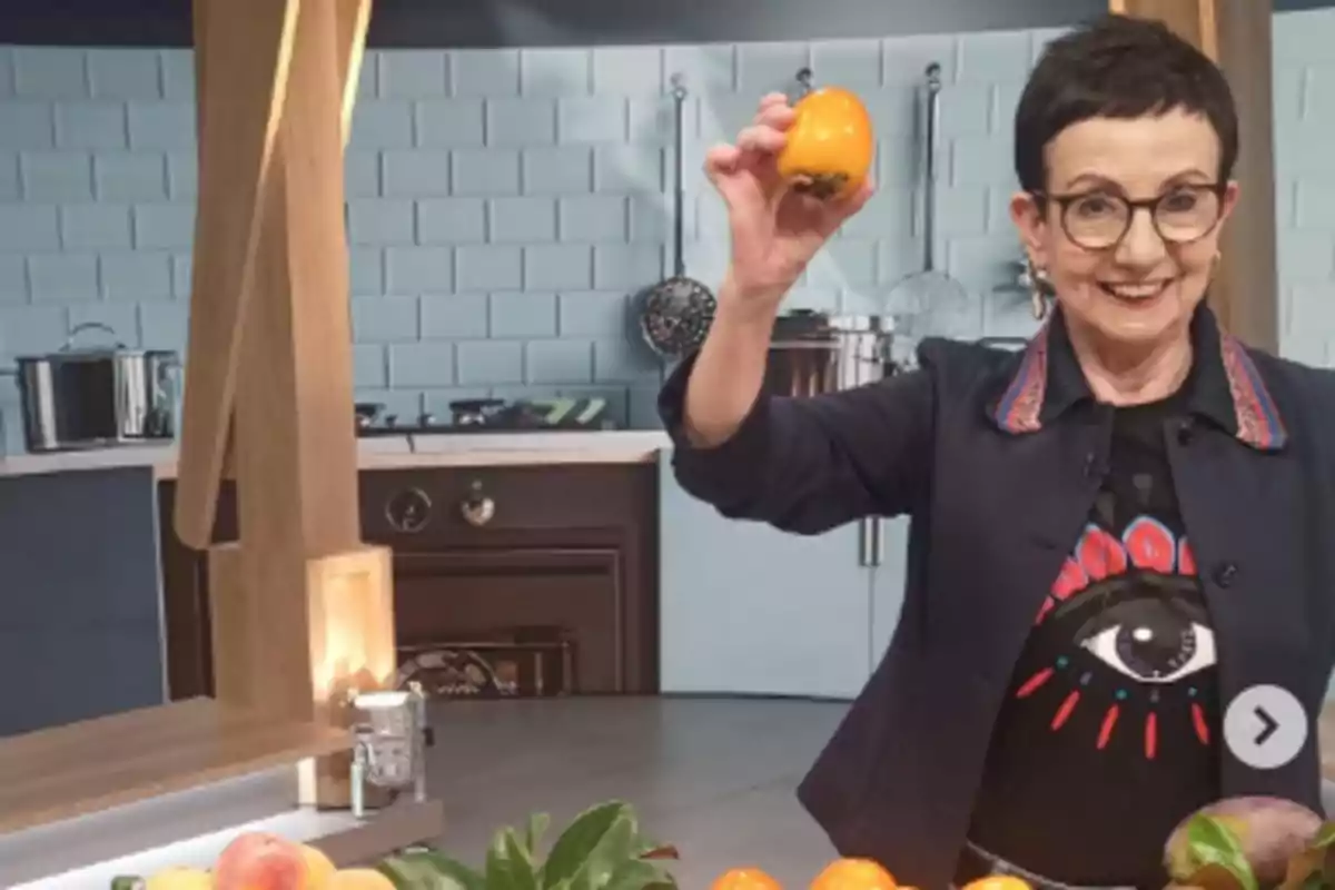 Una persona sonriente en una cocina moderna sostiene una fruta naranja mientras está rodeada de más frutas sobre la mesa.