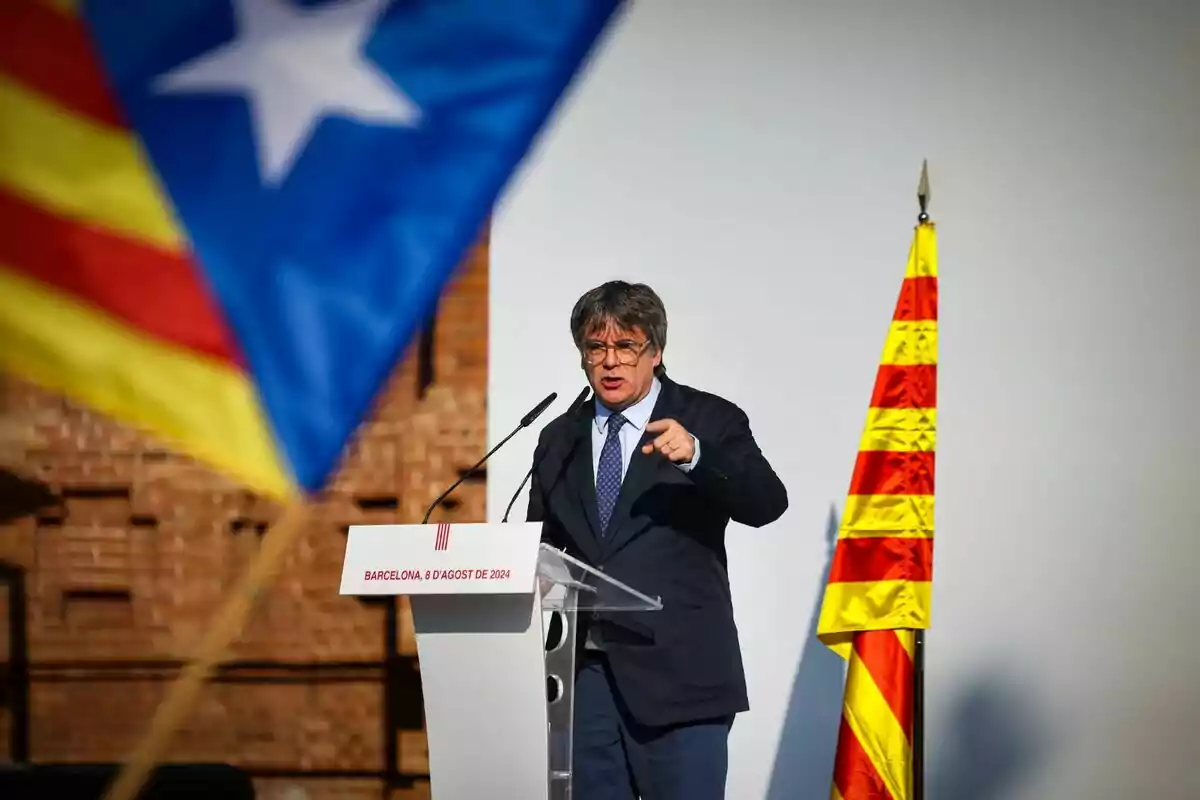 Un home parlant en un podi amb una bandera catalana i una bandera estelada al fons.