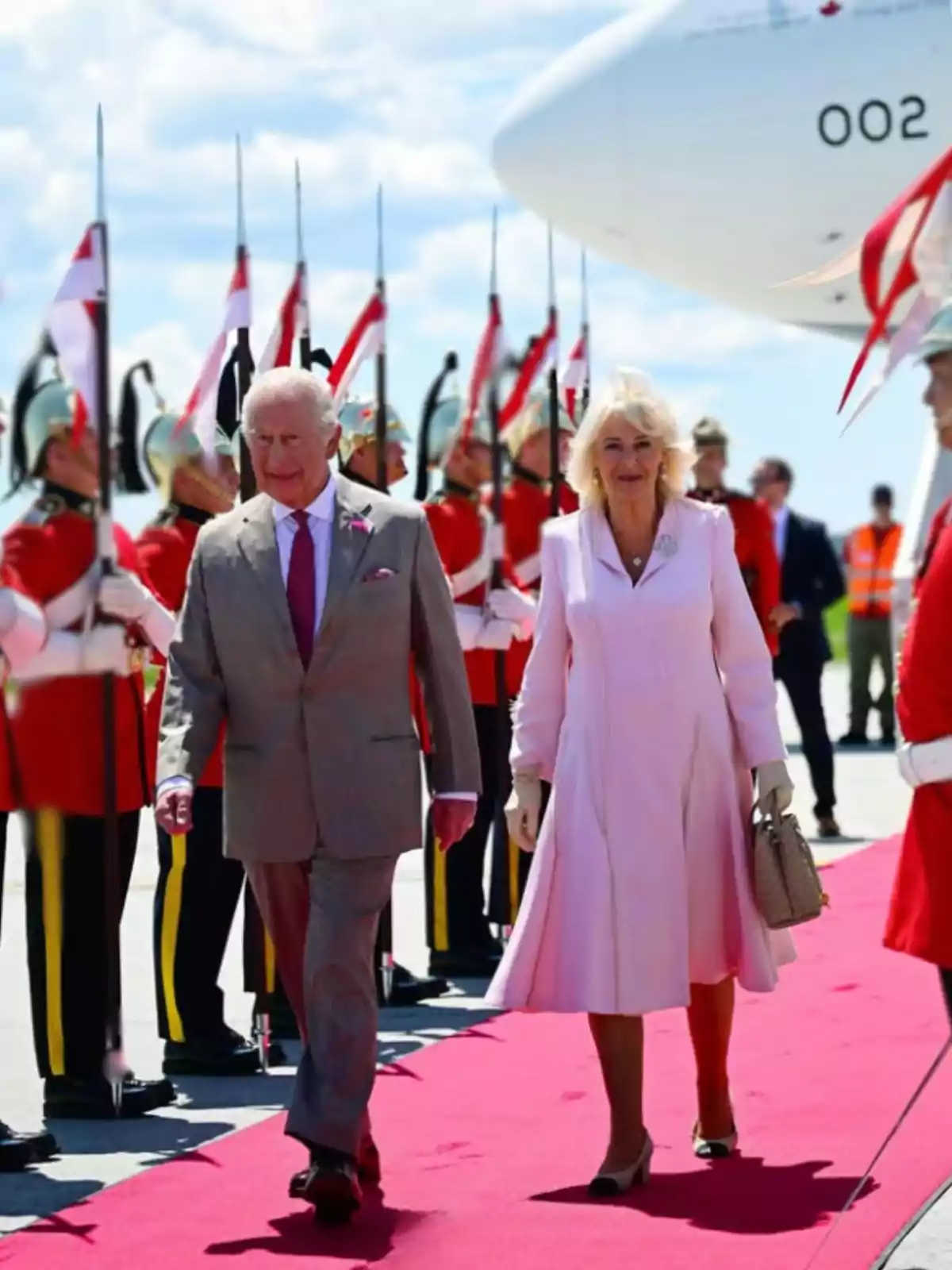 Una pareja elegantemente vestida camina por una alfombra roja mientras soldados con uniformes de gala y lanzas forman una guardia de honor junto a un avión blanco Una pareja elegantemente vestida camina por una alfombra roja mientras soldados con uniformes de gala y lanzas forman una guardia de honor junto a un avión blanco